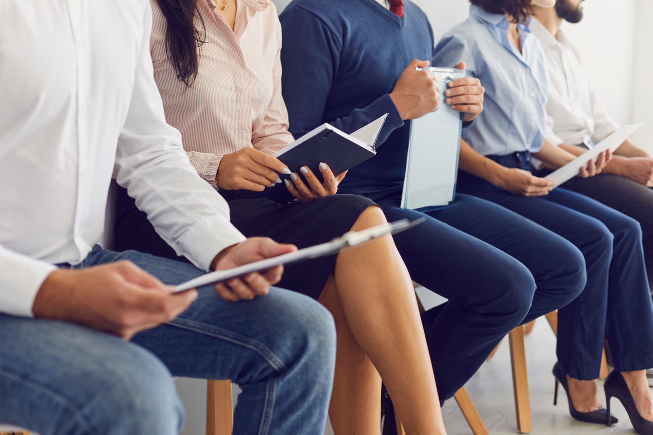 Cropped Image of People Sitting on Chairs in a Queue and Holding Paper in Their Hands.